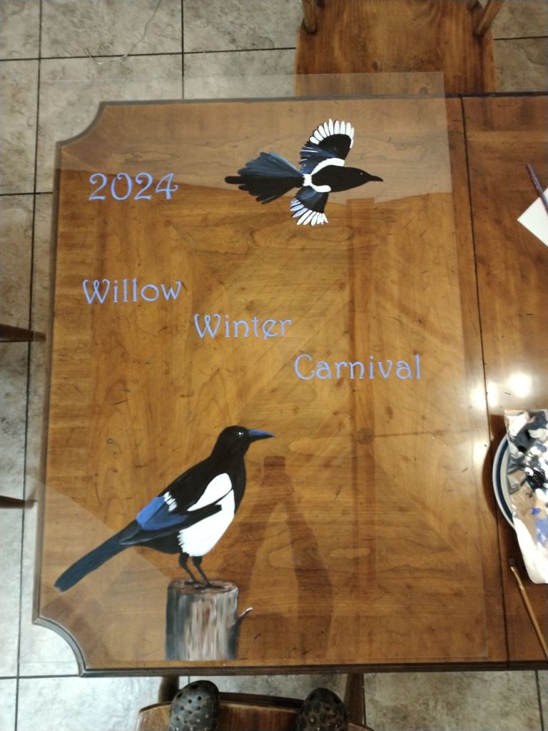 Painting of magpies on plexiglass.  2024 Willow Winter Carnival written across the center.